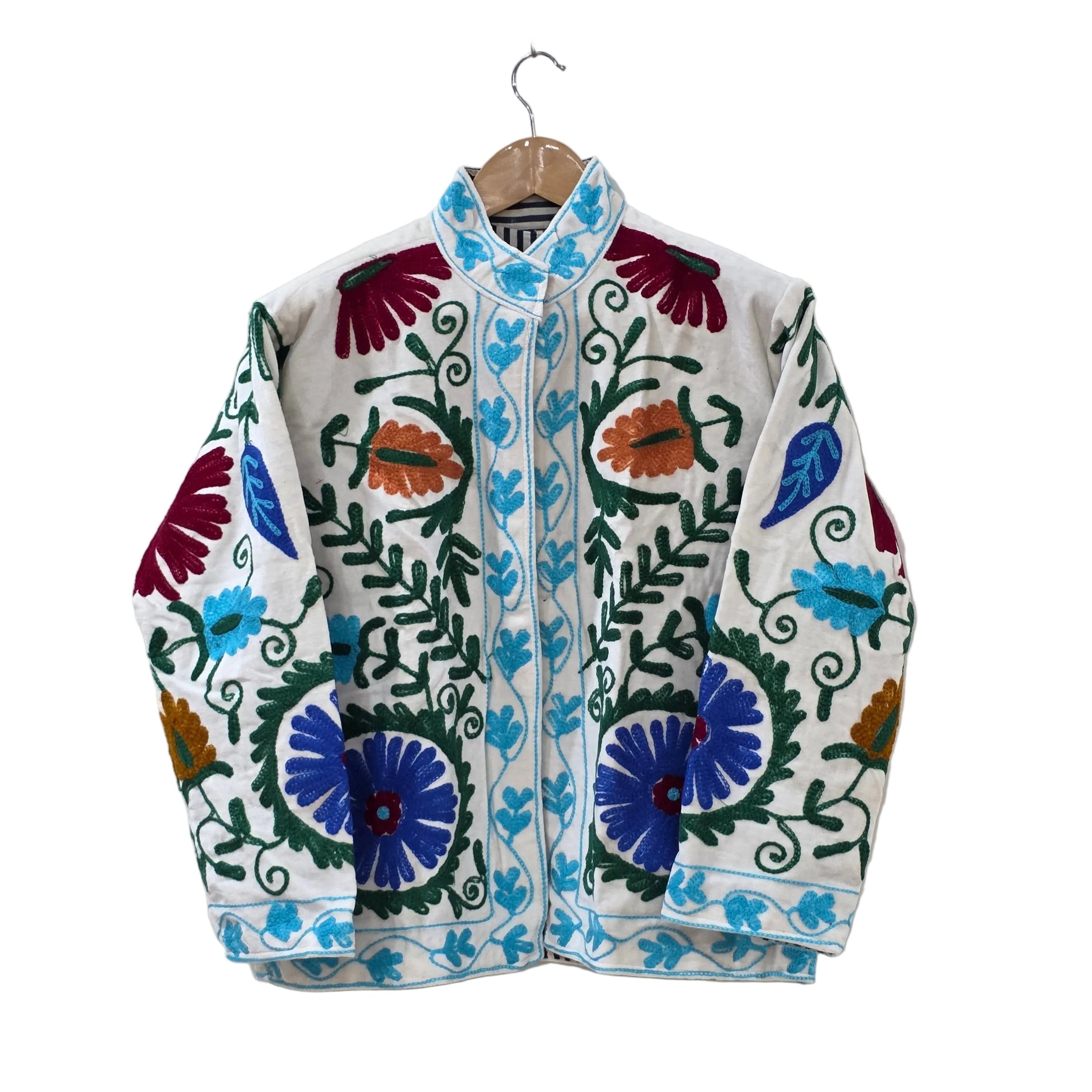 A cream white velvet bohemian jacket on a wooden hanger. It features traditional hand-stitched Suzani embroidery with large royal blue flowers near the hem, deep red blooms near the shoulders, winding dark green vines, and a contrasting bright turquoise blue embroidered border along the stand collar, front opening, and hem.