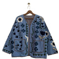 Light blue open-front TNT jacket with navy, white, and green embroidered floral, heart, and vine motifs, featuring two front patch pockets and striped trim, hanging on a wooden hanger.