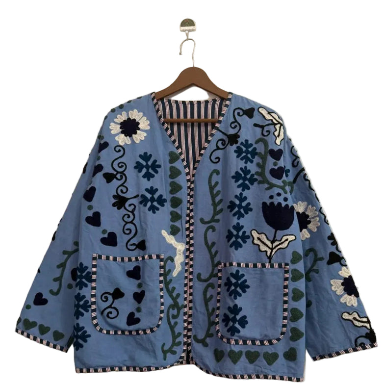 Light blue open-front TNT jacket with navy, white, and green embroidered floral, heart, and vine motifs, featuring two front patch pockets and striped trim, hanging on a wooden hanger.