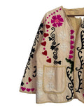 Front view of a cream-colored open-front TNT jacket featuring colorful embroidery including bright pink hearts, black vines, pink flowers, and white starbursts, finished with yellow and white striped piping and two front patch pockets.