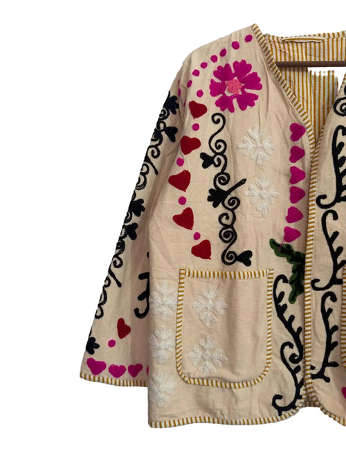 Front view of a cream-colored open-front TNT jacket featuring colorful embroidery including bright pink hearts, black vines, pink flowers, and white starbursts, finished with yellow and white striped piping and two front patch pockets.