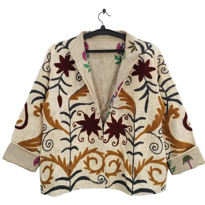 Front view of an open-style handmade cream textured cotton TNT short jacket featuring earthy rust brown leaves, dark vines, and maroon star-floral Suzani embroidery. The jacket is hanging on a black hanger against a white wall, slightly open with rolled cuffs to reveal a vibrant inner lining printed with magenta flowers and green palm trees.