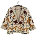 Front view of an open-style handmade cream textured cotton TNT short jacket featuring earthy rust brown leaves, dark vines, and maroon star-floral Suzani embroidery. The jacket is hanging on a black hanger against a white wall, slightly open with rolled cuffs to reveal a vibrant inner lining printed with magenta flowers and green palm trees.