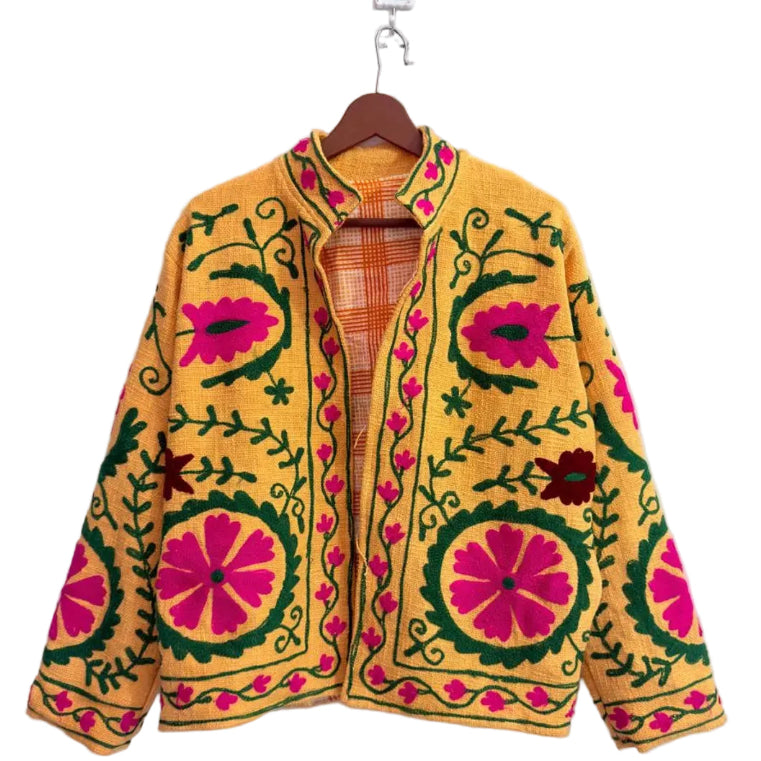 Close-up of a bright yellow textured TNT jacket featuring prominent crewel-style embroidery with large pink flowers and green vines, a stand collar, and an orange-and-white plaid inner lining.