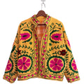 Close-up of a bright yellow textured TNT jacket featuring prominent crewel-style embroidery with large pink flowers and green vines, a stand collar, and an orange-and-white plaid inner lining.