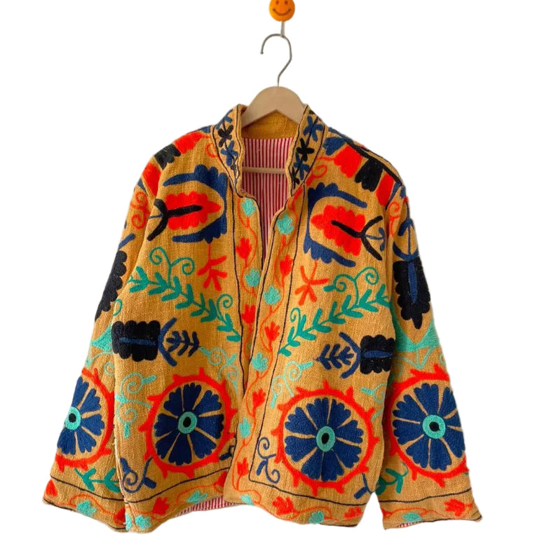 Front view of an open-style handmade mustard yellow textured cotton TNT short jacket featuring large navy blue, bright red, black, and turquoise Suzani floral and vine embroidery. The open front and mandarin collar reveal a red-and-white striped interior lining, hanging on a wooden hanger against a white wall as seen in the file