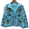 A vibrant sky blue hand-embroidered Suzani jacket with olive green, navy blue, and rust red floral botanical patterns hanging on a wooden hanger .