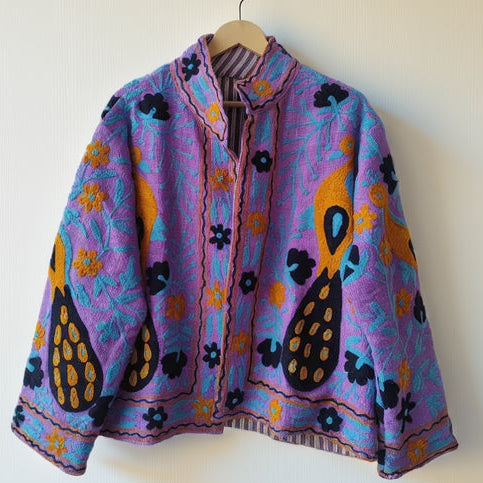 Bright purple open-front TNT jacket featuring turquoise vine embroidery, orange flowers, and bold black and orange paisley motifs, hanging on a wooden hanger against a white wall.