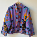 Bright purple open-front TNT jacket featuring turquoise vine embroidery, orange flowers, and bold black and orange paisley motifs, hanging on a wooden hanger against a white wall.