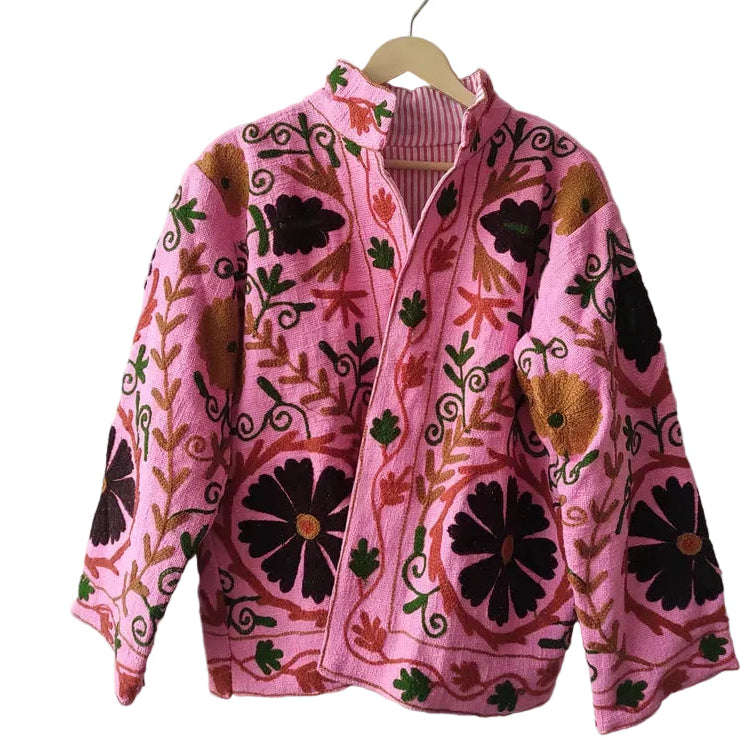 Light pink bohemian TNT jacket featuring oversized dark brown floral embroidery, green vine details, and an open front, hanging against a textured white wall.