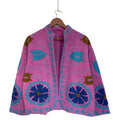 Bright pink open-front TNT jacket featuring large royal blue floral motifs, turquoise vine embroidery, and a stand collar, hanging on a wooden hanger against a white wall.