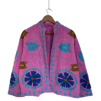 Bright pink open-front TNT jacket featuring large royal blue floral motifs, turquoise vine embroidery, and a stand collar, hanging on a wooden hanger against a white wall.