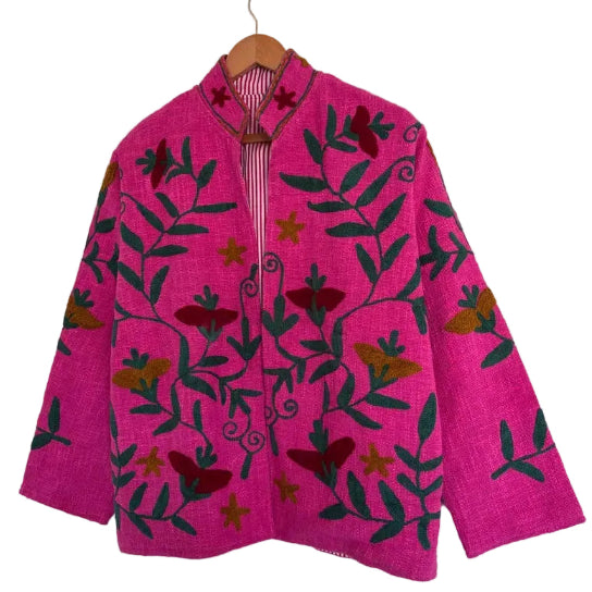 Front view of an open-style handmade hot pink textured cotton TNT short jacket featuring sprawling dark green vines and deep red and mustard yellow Suzani floral embroidery. The slightly open front and mandarin collar reveal a pink-and-white striped interior lining, hanging on a wooden hanger against a white wall .