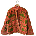 Back view of a coral pink jacket featuring intricate olive green embroidery with swirling vine, leaf, and bird motifs, hanging on a white wall.