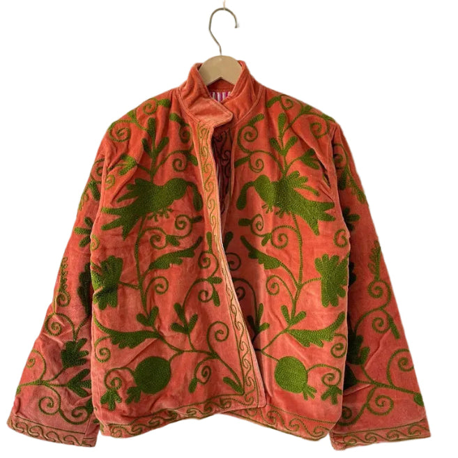 Back view of a coral pink jacket featuring intricate olive green embroidery with swirling vine, leaf, and bird motifs, hanging on a white wall.