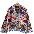 Cream open-front TNT jacket featuring vibrant pink, royal blue, purple, and earthy orange Suzani-style floral embroidery with a black and white striped inner lining, hanging on a wooden hanger.