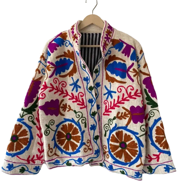Cream open-front TNT jacket featuring vibrant pink, royal blue, purple, and earthy orange Suzani-style floral embroidery with a black and white striped inner lining, hanging on a wooden hanger.